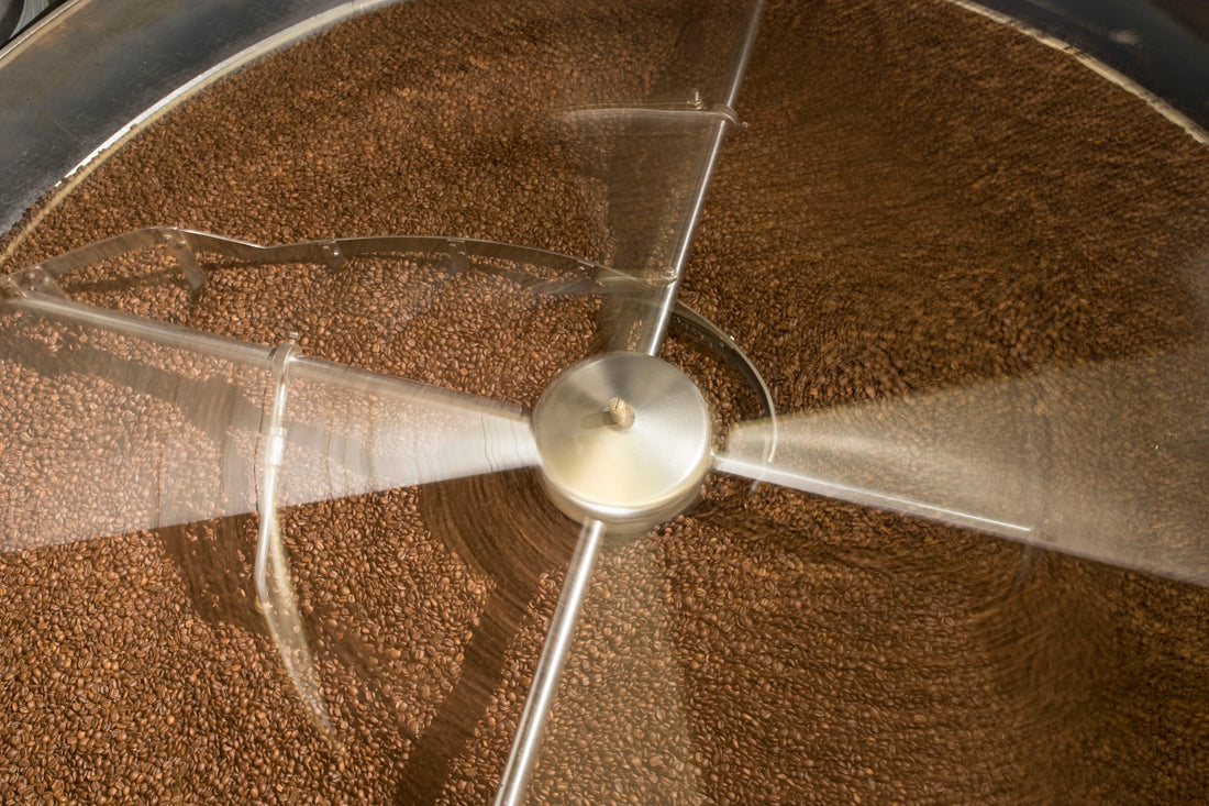 Nahaufnahme einer großen runden Kühlschale voller frisch gerösteter Kaffeebohnen, von oben fotografiert. Die metallenen Rührarme drehen sich sichtbar schnell und verteilen die Bohnen gleichmäßig zum Abkühlen.