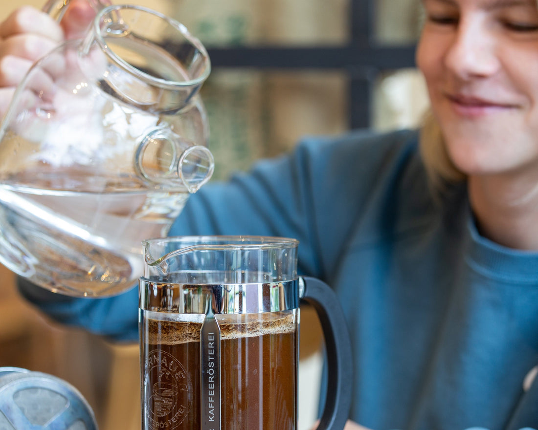  Person gießt Wasser in eine French Press mit Cold Brew Kaffee der Murnauer Kaffeerösterei und lächelt bei der Zubereitung, Cold Brew Kaffee selber machen: Rezept, Methoden und Tipps für zu Hause