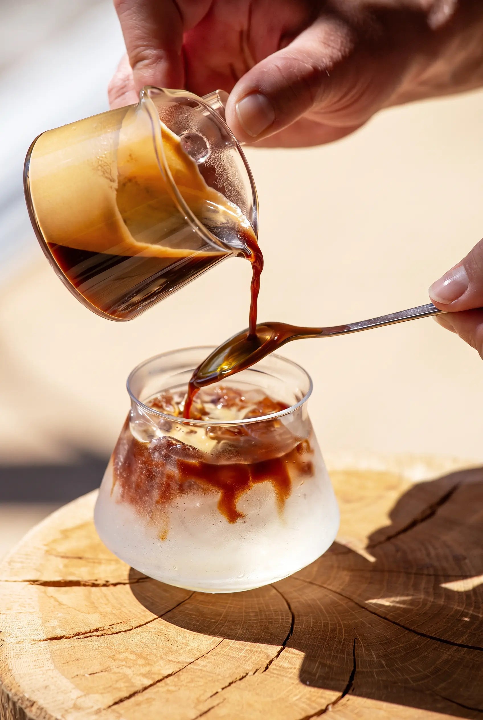 Barista giesst Espresso für Espresso Tonic über Eiswürfel ins bauchige Glas auf Holz, mit Löffel geführt.