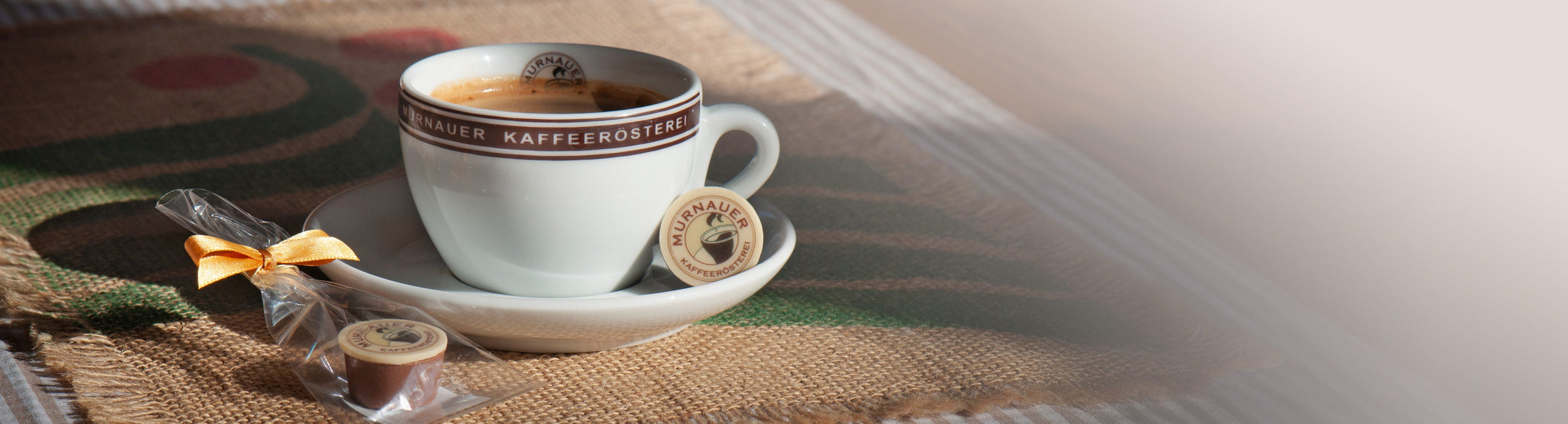 Kaffeetasse der Murnauer Kaffeerösterei auf Jutesack mit eingepackter Praline daneben als Banner für das Kaffee-Gesamtsortiment
