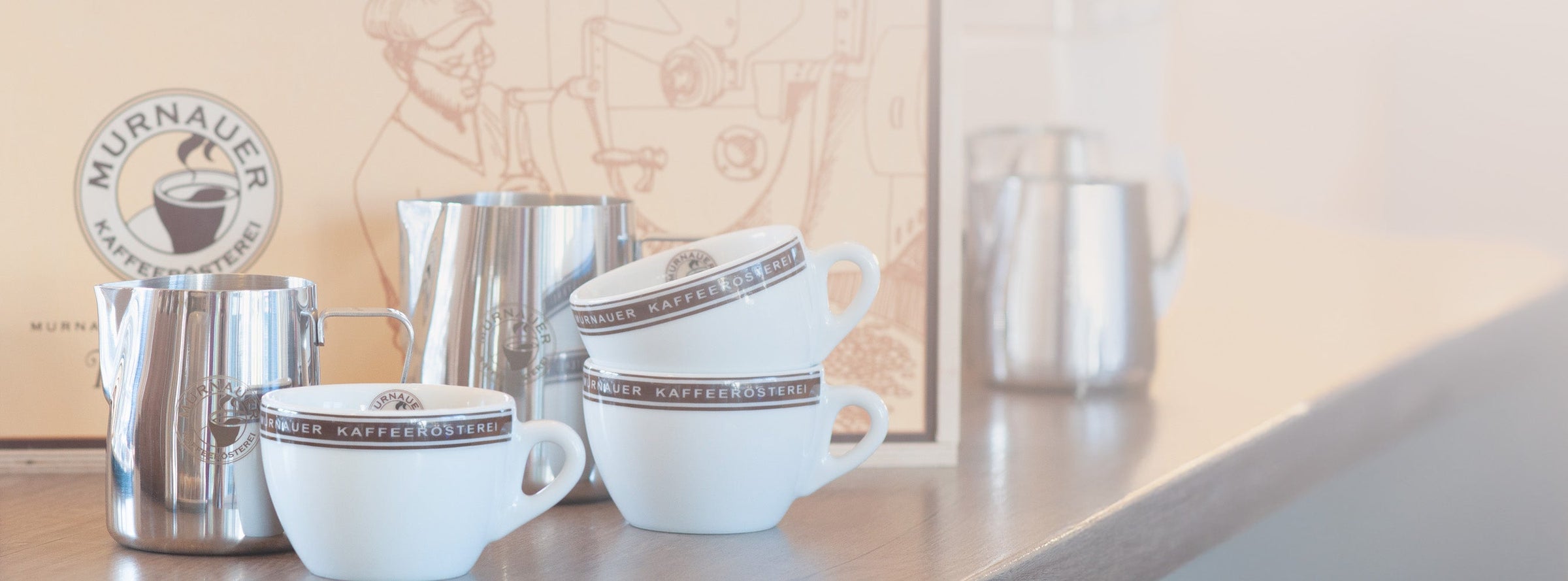 Stapel aus Espresso- und Cappuccinotassen sowie Edelstahl-Milchkännchen mit Logo der Murnauer Kaffeerösterei auf einer Theke
