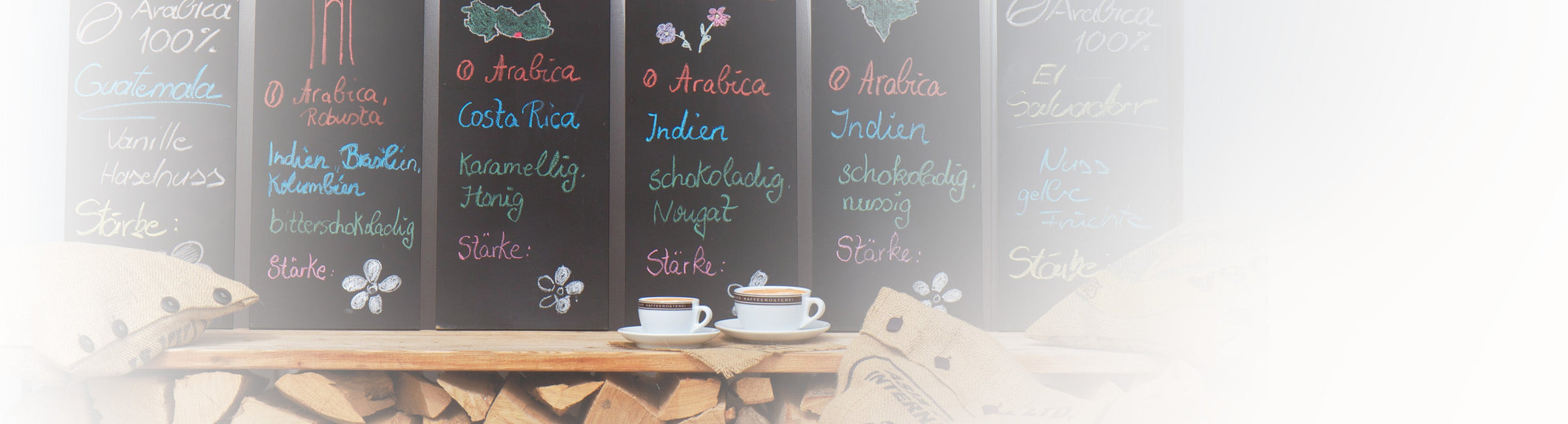 Schwarze Kreidetafeln mit Kaffeemischungen und zwei Espressotassen vor Holz und Jutesäcken als Banner für Kaffee-Entdeckungsreisen