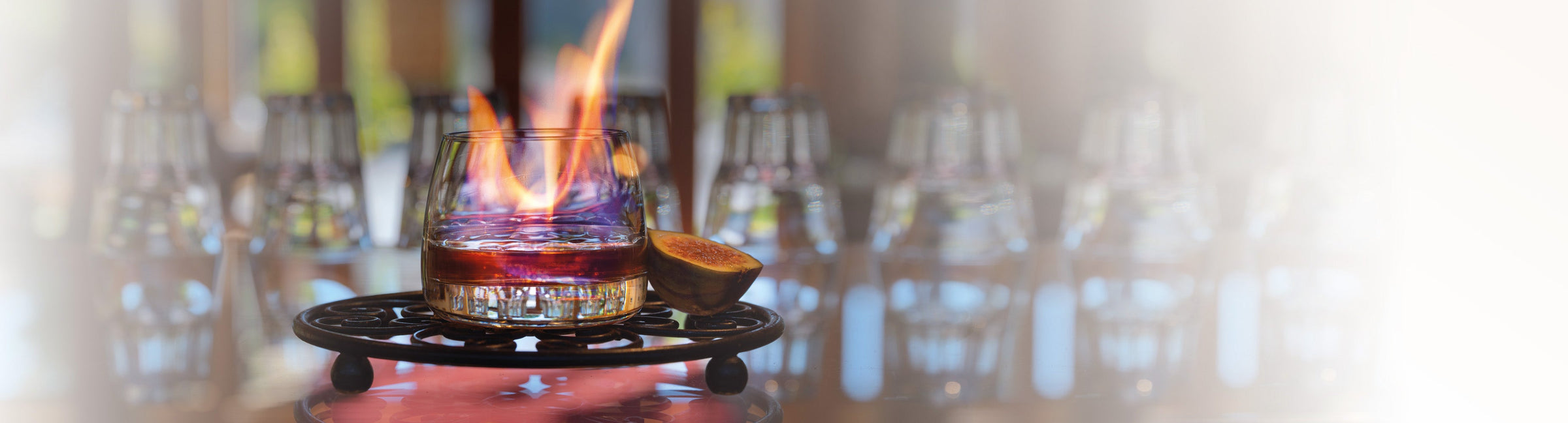 Brennender Spirituosen-Drink im Glas auf Metalluntersetzer mit Feige im Vordergrund, unscharfe Gläser im Hintergrund als Banner für Spirituosen