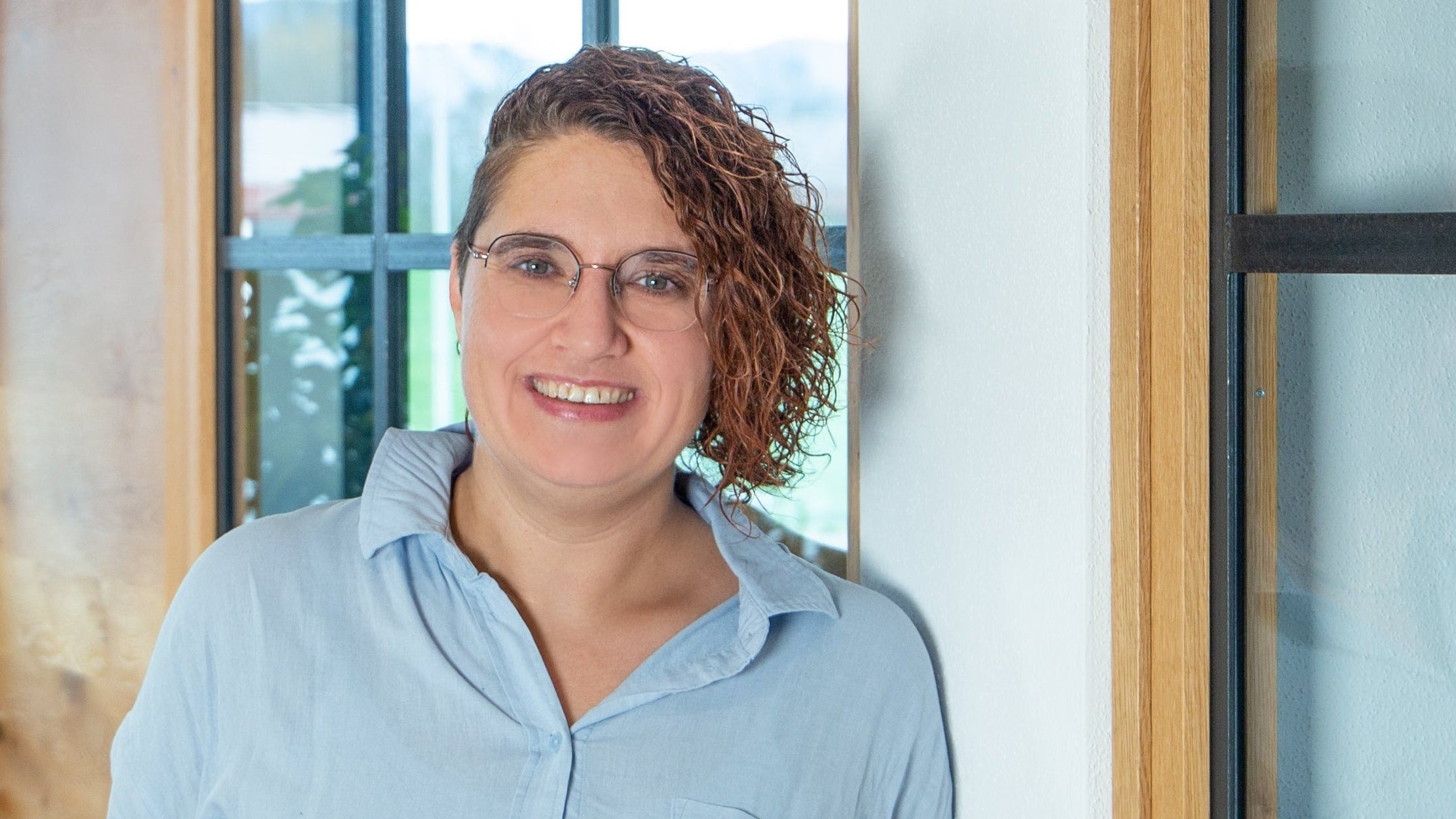 Mitarbeiterin im Marketing der Murnauer Kaffeerösterei in hellblauer Bluse lehnt lächelnd im Büroflur an einer Glaswand, Andrea Nebl