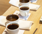 Kaffee-Kurs Brühen und Mahlen der Kaffee Academy im Murnauer Kaffeehaus: Kaffeeverkostung mit vier Cupping Tassen Filterkaffee, Servietten und Cupping-Löffel auf Holztisch