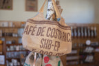 Upcycled Tragetasche aus bedrucktem Kaffeesack im Kaffeehaus, hängt an Birkenstamm, Regale mit Kaffeewaren im Hintergrund