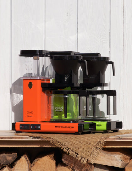Orange und grüne Moccamaster Filterkaffeemaschine mit zwei Glaskannen auf Holzbrettern vor weißer Holzwand in der Sonne
