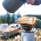 Outdoor-Kaffee mit Emaille-Tasse und Handfilter auf Bergwiese, Hand gießt heißes Wasser aus Campingtopf in den Kaffeefilter - Janna Kamphoff