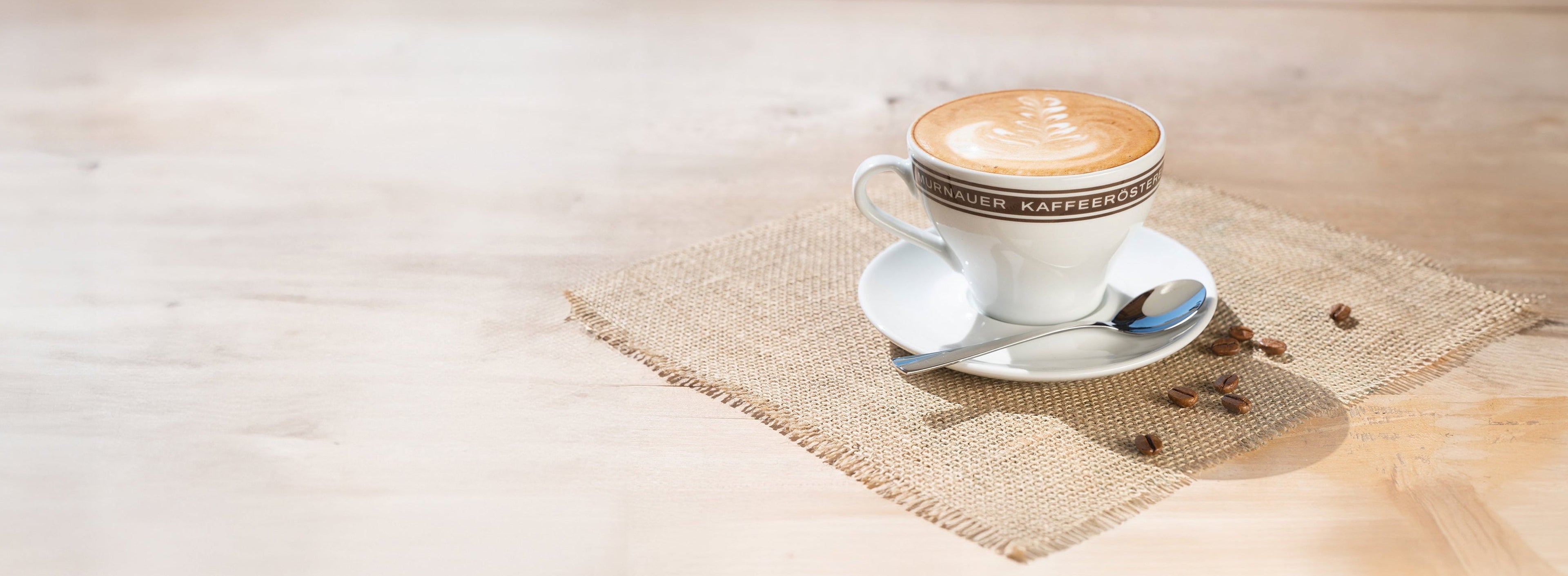 Frische Cappuccino-Tasse mit Latte Art der Murnauer Kaffeerösterei auf Untertasse mit Löffel und Juteserviette auf Holztisch