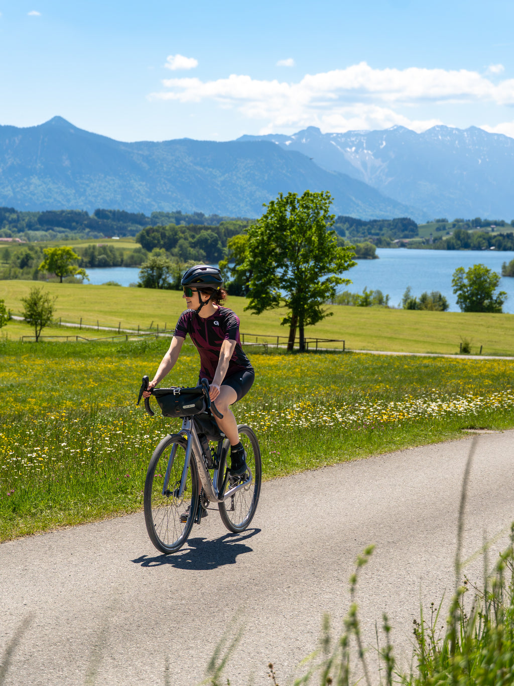 Radfahrerin auf Rennrad fährt auf Landstraße am See vor bayerischen Alpen unter blauem Himmel, Coffee Ride Feeling - Bild: Janna Kamphoff