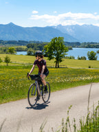 Radfahrerin auf Rennrad fährt auf Landstraße am See vor bayerischen Alpen unter blauem Himmel, Coffee Ride Feeling - Bild: Janna Kamphoff