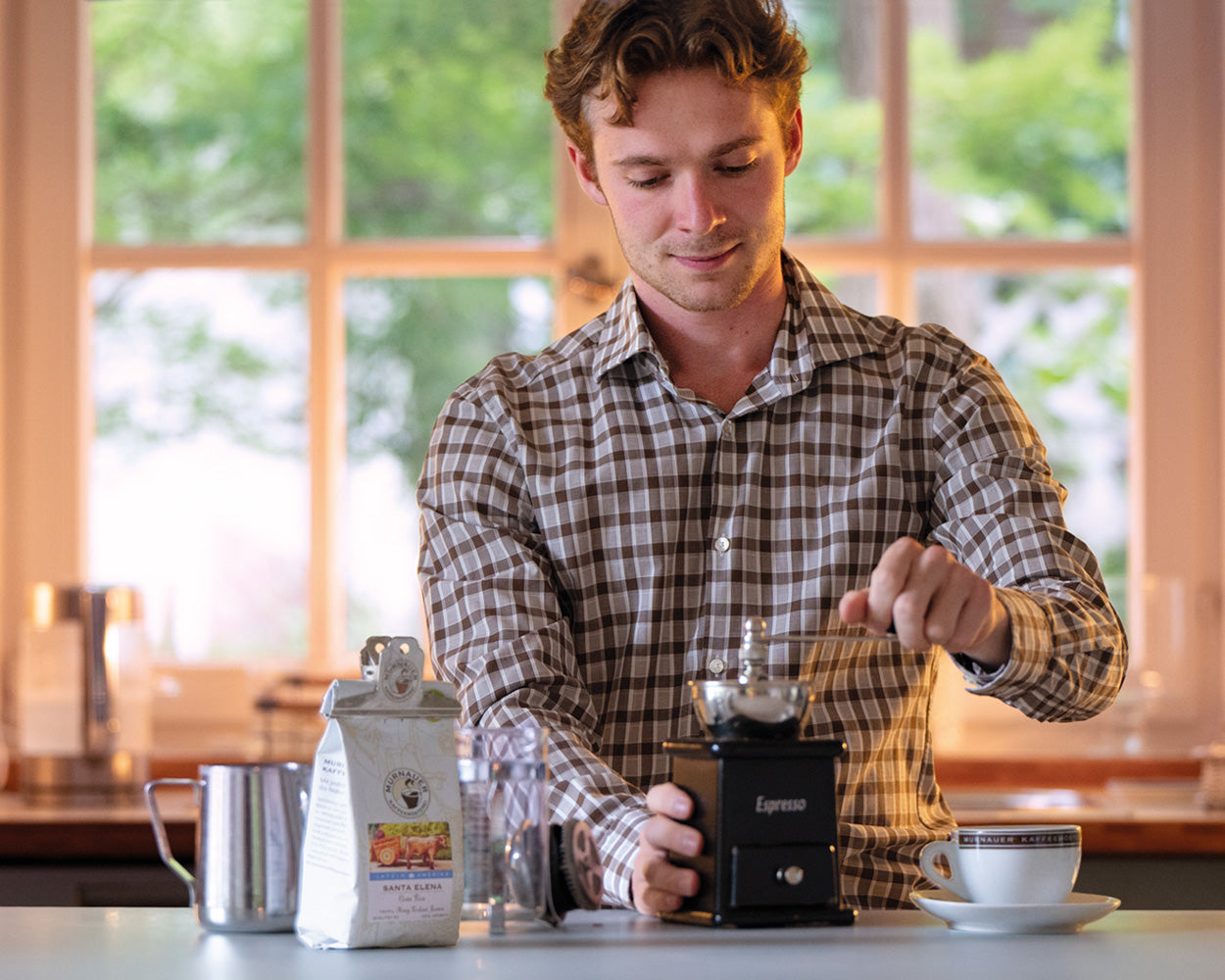 Junger Mann mahlt Kaffee mit einer Handmühle, daneben Santa Elena Kaffeepackung und Cappuccinotasse der Murnauer Kaffeerösterei auf der Arbeitsplatte