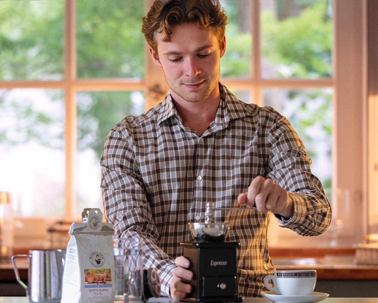 Junger Mann mahlt Kaffee mit schwarzer Handmühle in heller Küche, Santa Elena Kaffeepackung und Espressotasse der Murnauer Kaffeerösterei stehen daneben