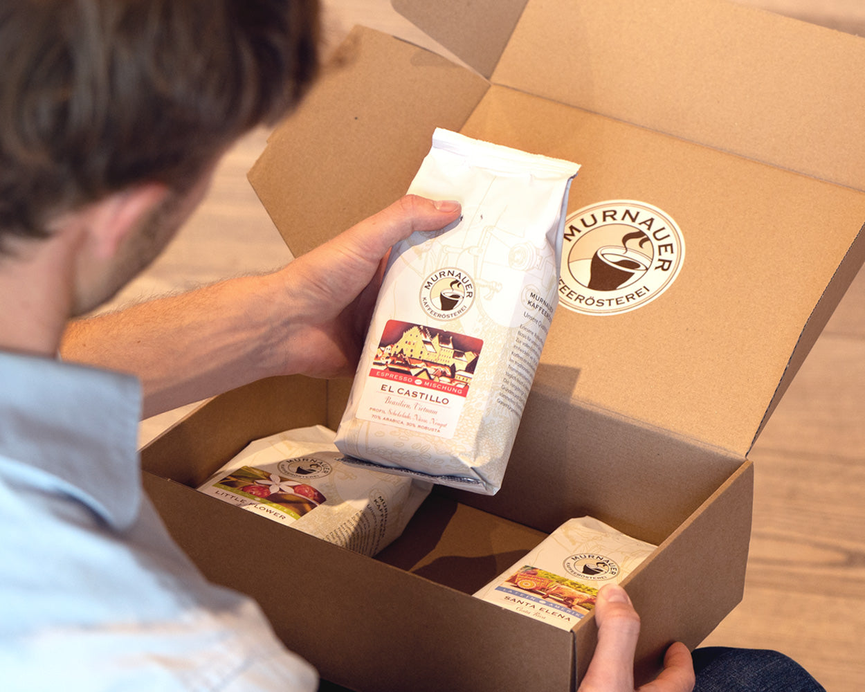Person öffnet Versandkarton der Murnauer Kaffeerösterei und hält El Castillo Kaffeepackung in der Hand, weitere Kaffeetüten liegen im Paket