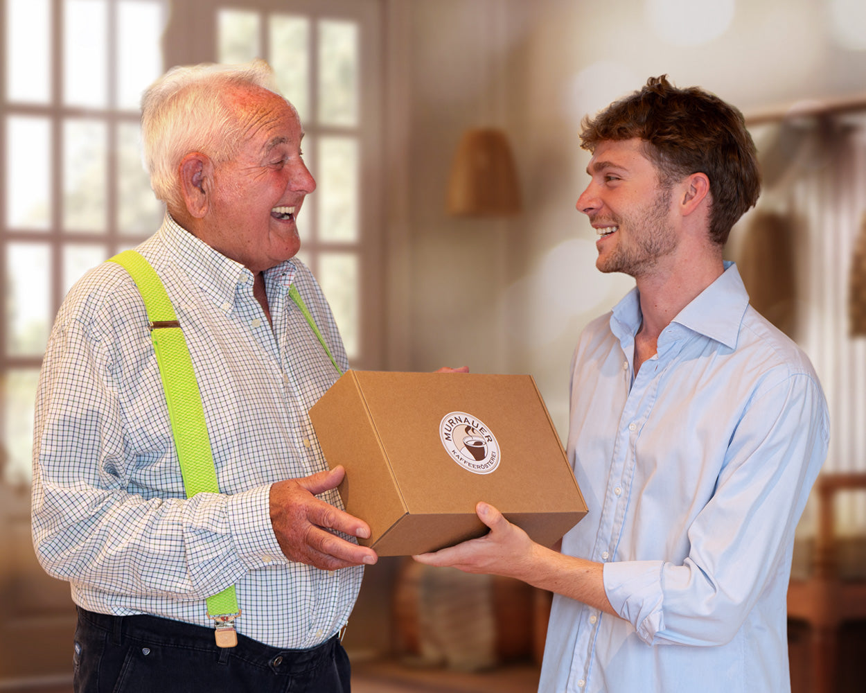 Älterer Mann gibt lachend eine Geschenkbox der Murnauer Kaffeerösterei von jüngerem Mann, beide stehen im warm beleuchteten Raum