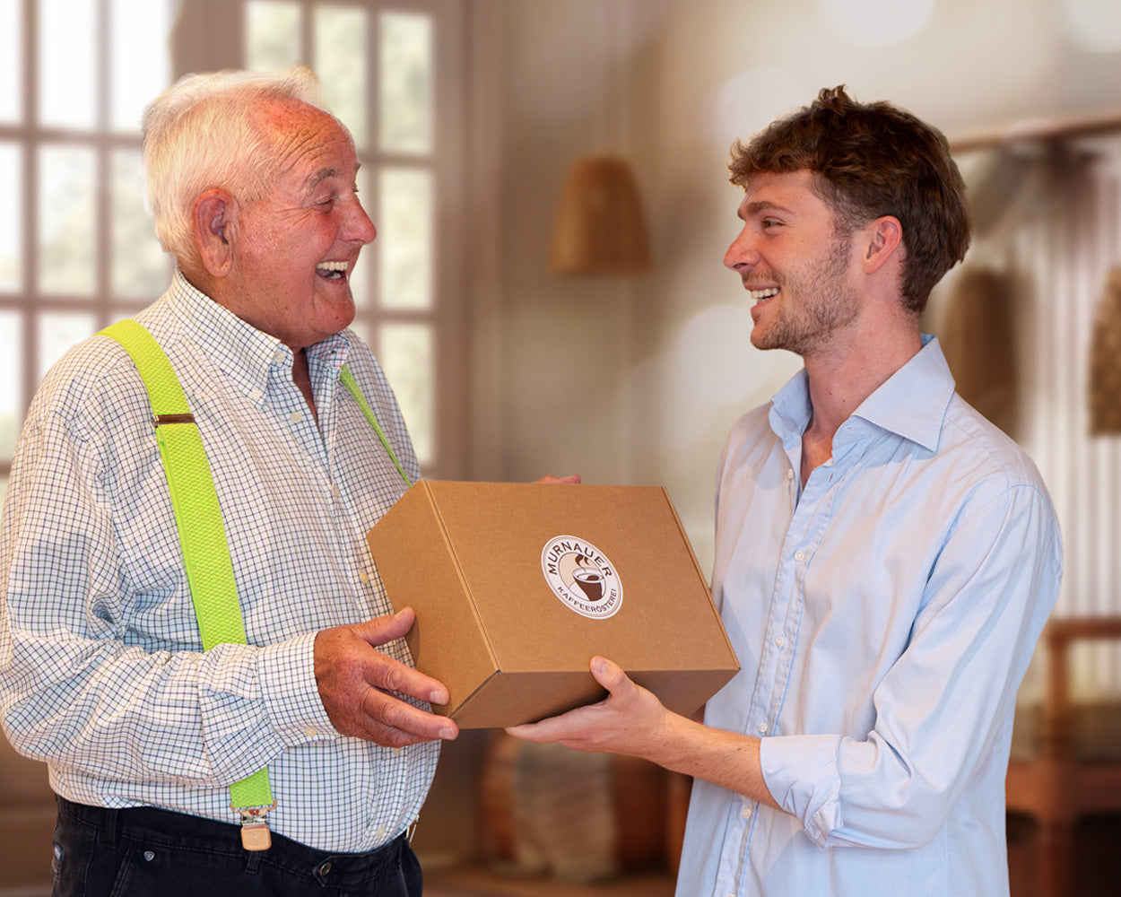 Älterer Mann gibt lachend eine Geschenkbox der Murnauer Kaffeerösterei von jüngerem Mann, beide stehen im warm beleuchteten Raum