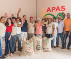 Gruppe von Kaffeebäuerinnen und -bauern der APAS Kooperative in Brasilien vor Kaffeesäcken und APAS Logo, alle lachen und winken