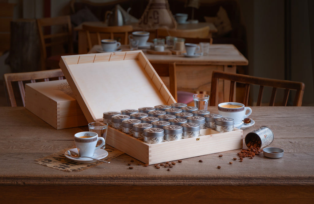Kaffee-Adventskalender 2026 der Murnauer Kaffeerösterei in geöffneter Holzbox auf Holztisch, daneben Cappuccino-Tasse, Glas Wasser und verstreute Kaffeebohnen in gemütlicher Atmosphäre eines Cafès