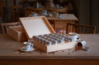 Kaffee-Adventskalender 2026 der Murnauer Kaffeerösterei in geöffneter Holzbox auf Holztisch, daneben Cappuccino-Tasse, Glas Wasser und verstreute Kaffeebohnen in gemütlicher Atmosphäre eines Cafès