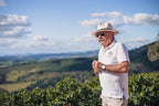 Produzent auf der Fazenda Rainha Brasilien Kaffeeplantage mit Hut und Sonnenbrille steht vor grünem Hügelland unter blauem Himmel