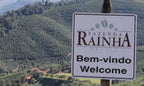 Schild mit Aufschrift Fazenda Rainha Brasil, Bem-vindo Welcome, vor grünen Hügeln einer Kaffeeplantage in Brasilien