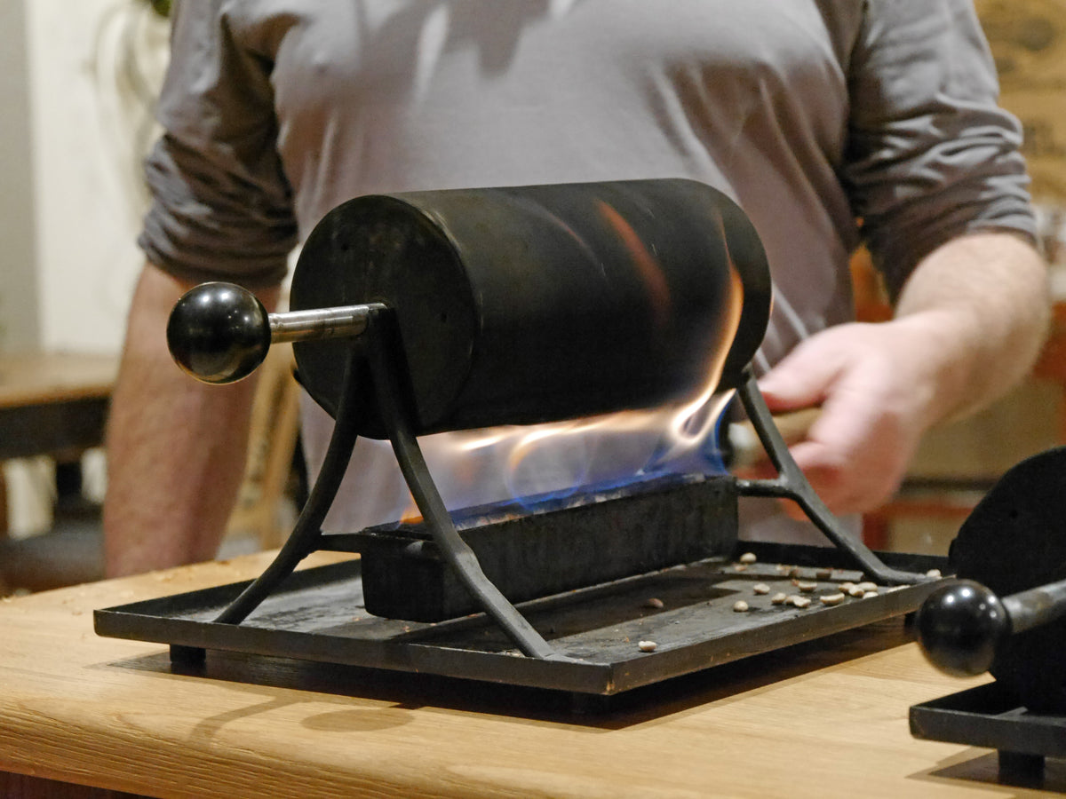 Kaffeerösten Kurs mit Inhaber Thomas Eckel in der Kaffee Academy im Murnauer Kaffeehaus, Handröster über Gasflamme mit röstenden Bohnen auf Holztheke