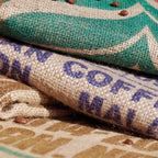 Gestapelte Jute Kaffeesäcke mit blauem Aufdruck und grünem Design, dazwischen verstreute Kaffeebohnen als natürliche Deko, Motive symbolisch - Auswahl erfolgt willkrlich je nach Verfügbarkeit