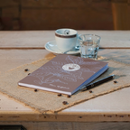 Braunes Kaffeetagebuch der Murnauer Kaffeerösterei mit Stift auf Jutesack, dahinter Espressotasse und Wasserglas auf Holztisch