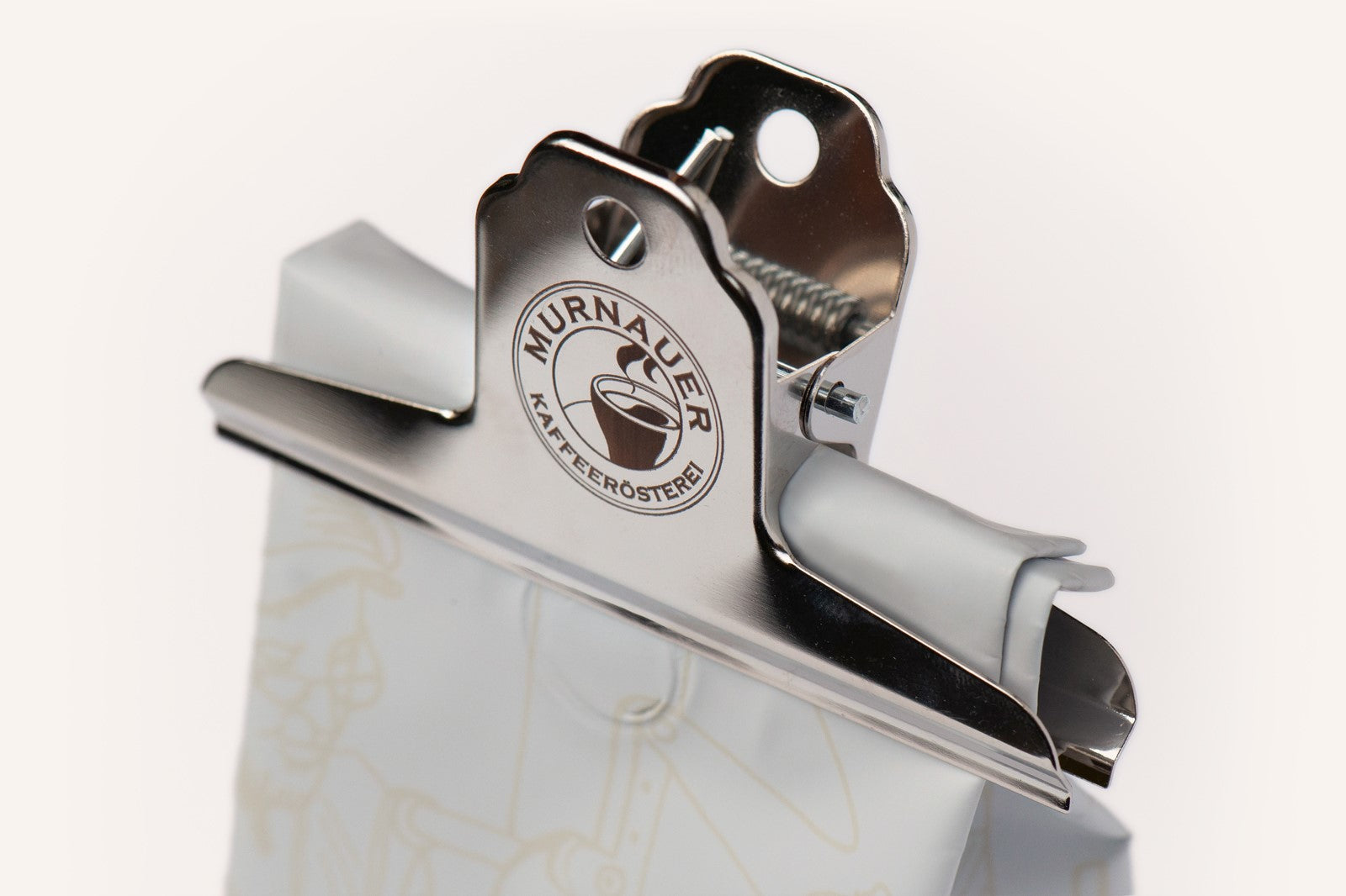 Metall Kaffeetüten Clip mit Logo der Murnauer Kaffeerösterei verschließt eine 250 g Kaffeetüte luftdicht von vorn, zeigt Größenverhältnis von 250g Tüte und dem Clip