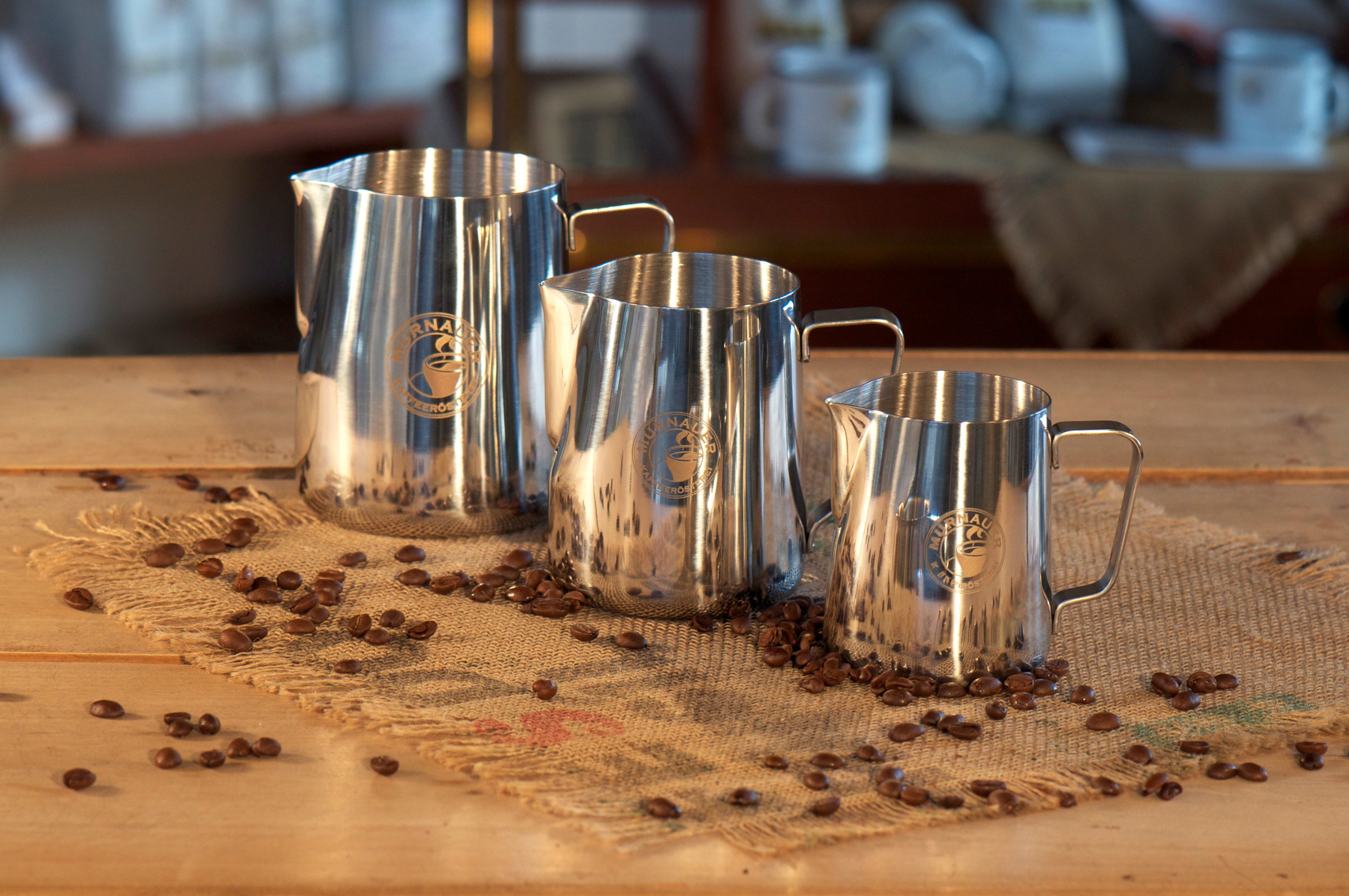 Drei Edelstahl Milchkännchen der Murnauer Kaffeerösterei in verschiedenen Größen stehen auf Jutesack mit verstreuten Kaffeebohnen, 350 ml, 590 ml, 950 ml