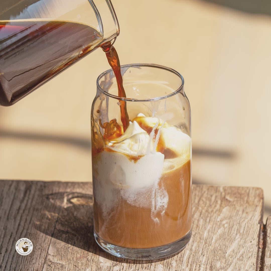 Eiskaffee Zubereitung - Ausschnitt: Hand gibt Zutaten Vanilleeis, Cold brew und Sahne in hohes Glas auf Holztisch im Sonnenlicht