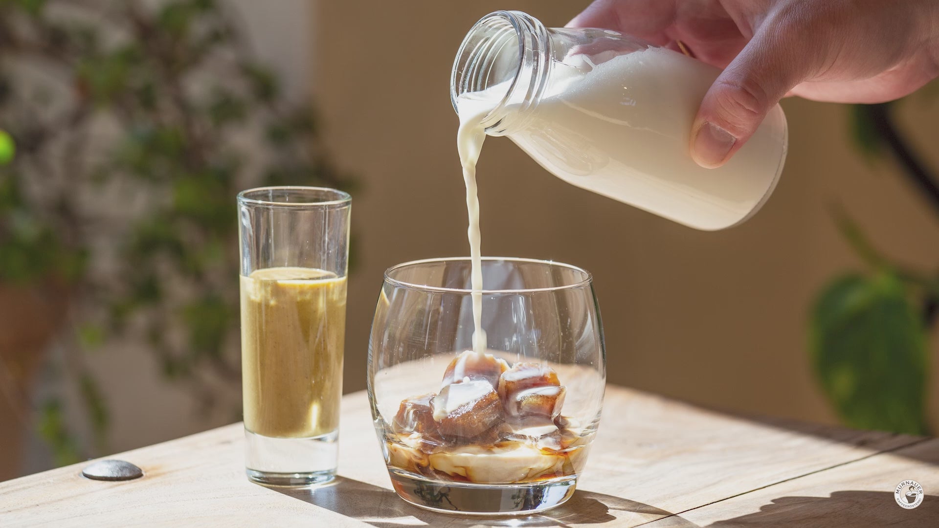 Milch wird in Glas mit Kaffee-Eiswürfeln für Pistazien Spanish Latte gegossen, daneben hohes Glas mit cremiger Pistazienmischung, Rezept der Murnauer Kaffeerösterei - VIDEO