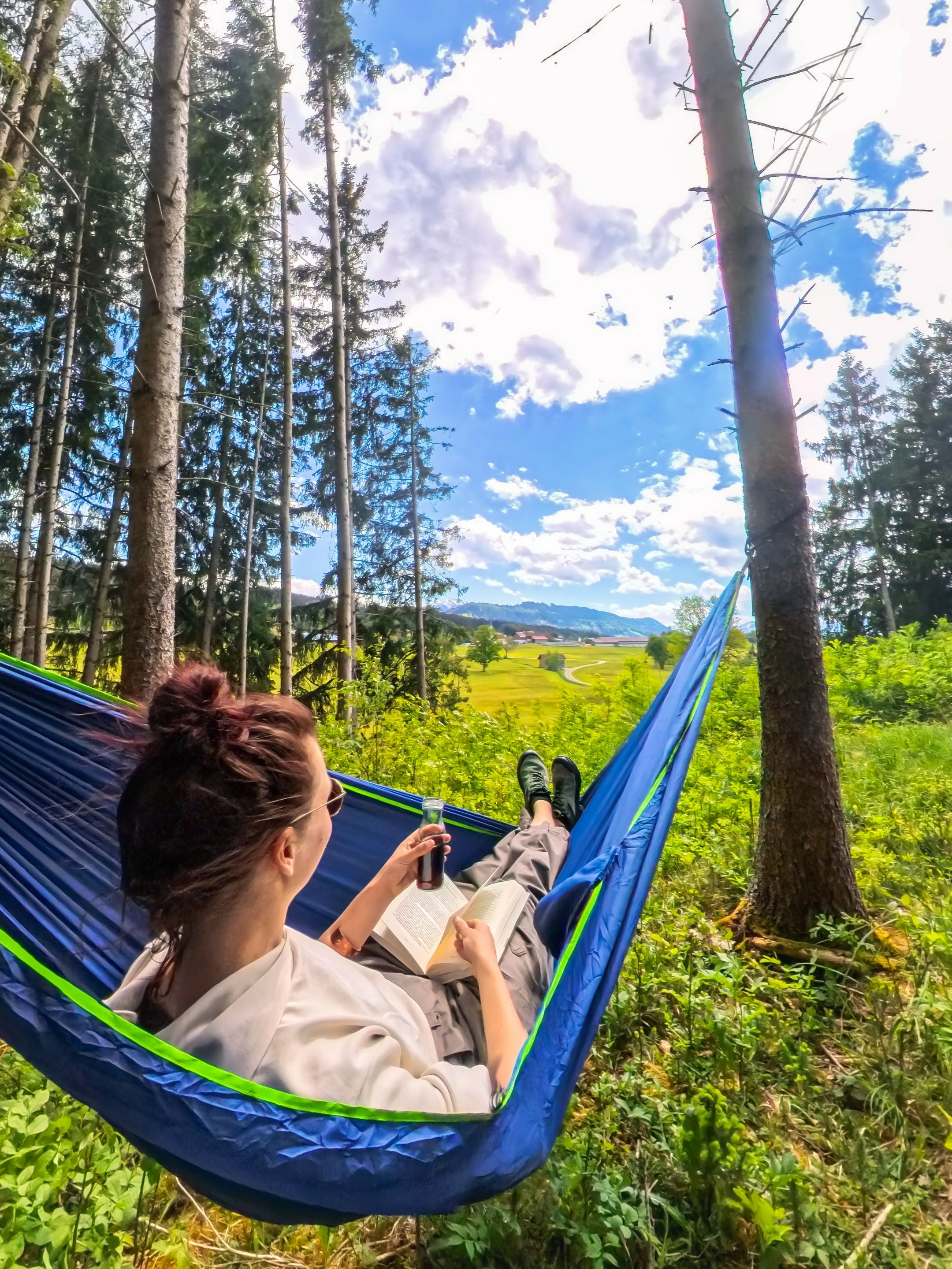 Frau entspannt mit Kaffeelust Unterwegs Kaffee in blauer Hängematte im Wald mit Blick auf sonnige Wiesenlandschaft, Janna Kamphoff
