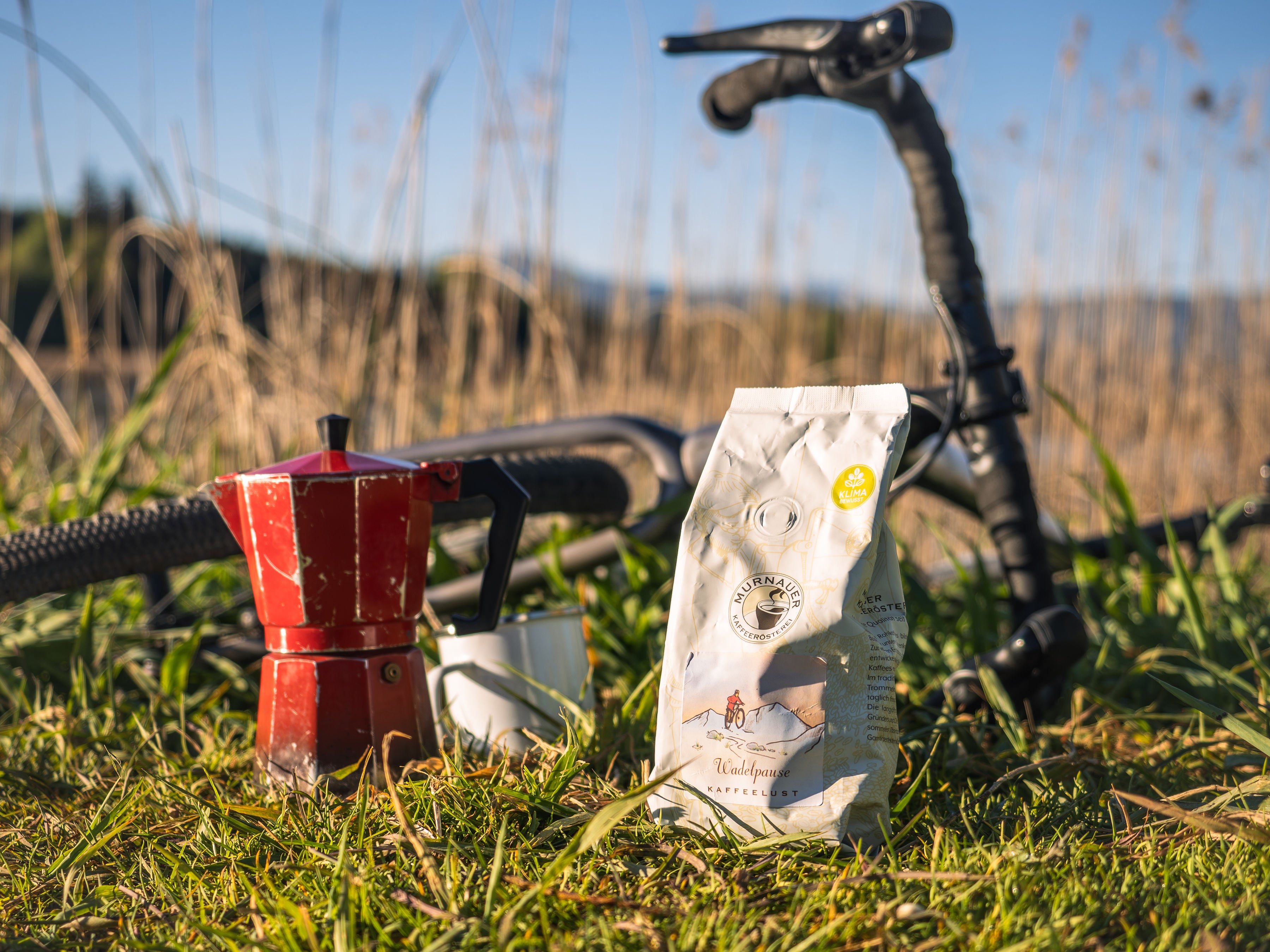 Kaffeelust Wadelpause Kaffee im Beutel mit roter Mokkakanne und Tasse auf Wiese neben Fahrradlenker in der Sonne