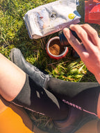 Radfahrerin sitzt in Wiese, füllt Mokkakanne mit gemahlenem Kaffeelust Wadelpause Kaffee neben Packung und Radsportsocken, Bild: Janna Kamphoff