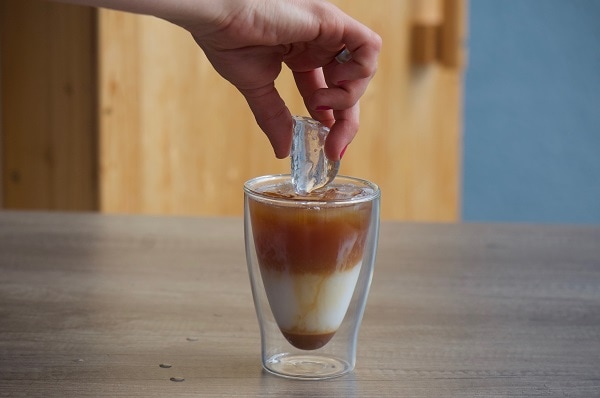 Glas befüllt mit Sirup, Milchschaum und Kaffee, dazu eine Hand, die einen Eiswürfel ins Glas gibt von oben herab