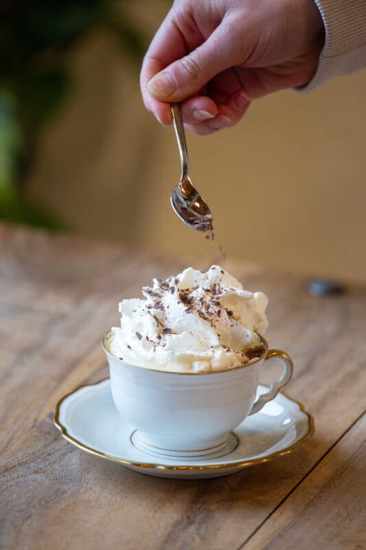 Zubereiteter Pharisäer-Kaffee mit Sahnehaube in Tasse und Person streut Schokostreusel auf Sahne