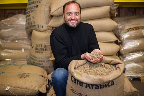 lachender Mann kniet vor Kaffeesack und haelt zwei gefuellte Haende voller roher Kaffeebohnen in die Kamera