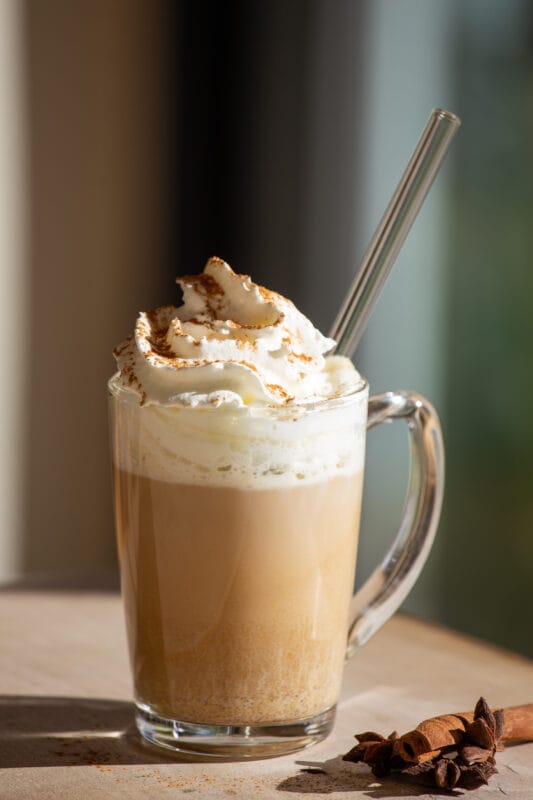 Pumpkin spice latte mit sahne und zimt bestreut, sternanis und zimtstangen auf holztisch von der sonne angestrahlt