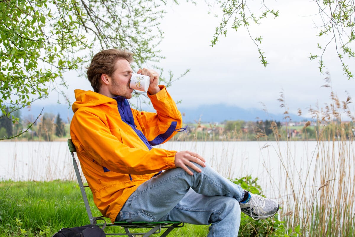draußen kaffee trinken: mann trinkt sitzt inm stuhl draussen am see einen kaffee aus einer emailletasse