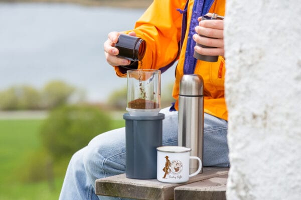 Outdoor-Kaffeezubereitung mit Handfilter: Person in orangefarbener Jacke bereitet frisch gemahlenen Kaffee mit einem Handfilter neben Thermoskanne und Emaille-Tasse von Murnauer Kaffeerösterei zu.
